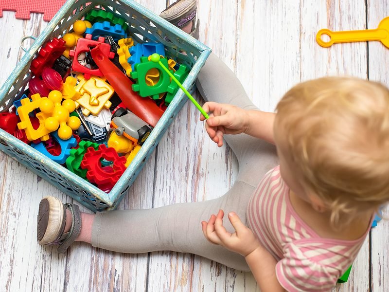 Small Box Filled With Childrens Small Toys For Education. View F Small box filled with childrens small toys for education. View from top. little kid sits near box of developing toys. Toys liners are multicolored. Shapes. Color Recognition Shape Toys with Colorful Sorter